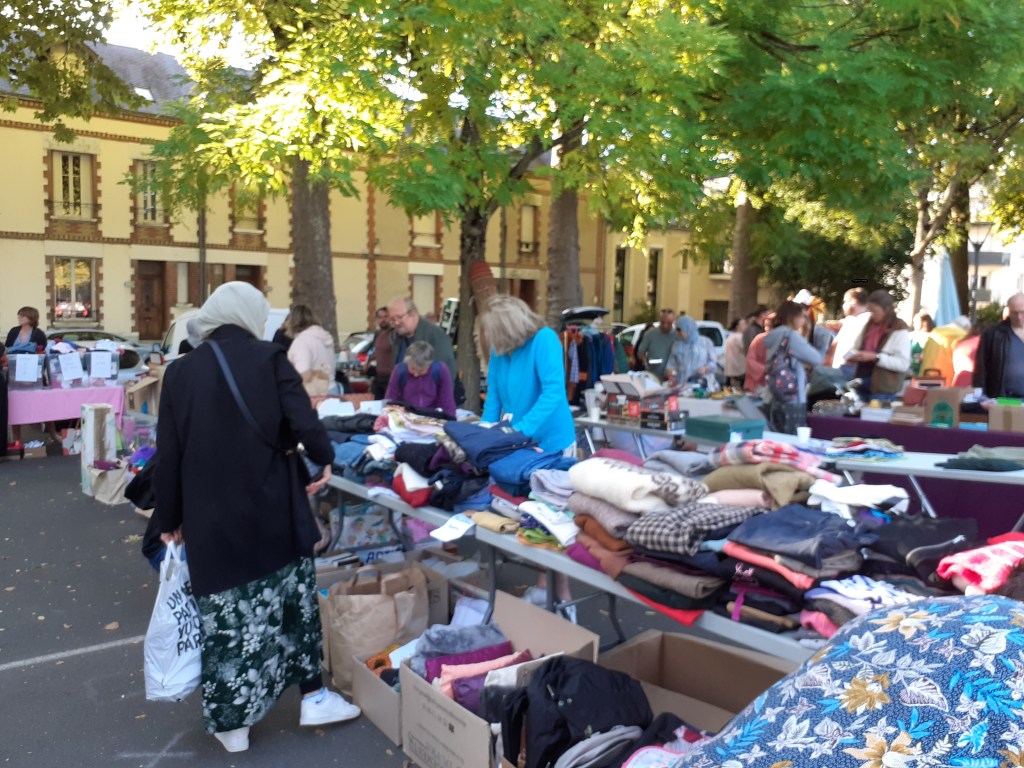 Marché aux puces animé avec des tables chargées de vêtements et d'objets divers, des visiteurs explorant les étals sous un ciel bleu.